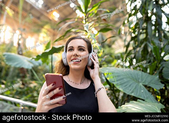 Happy beautiful young woman listening music through wireless headphones in front of plants