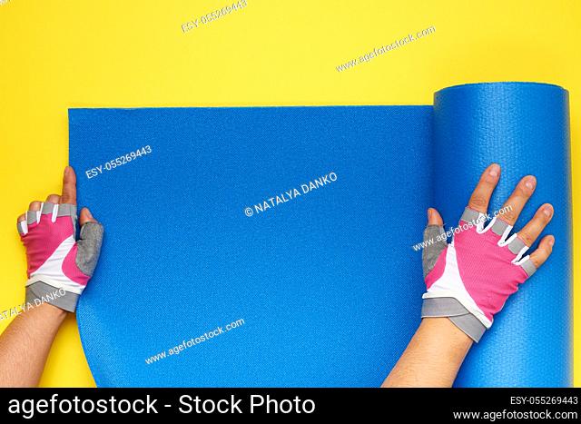 two female hands in sports gloves unfold a blue yoga mat, top view, yellow background