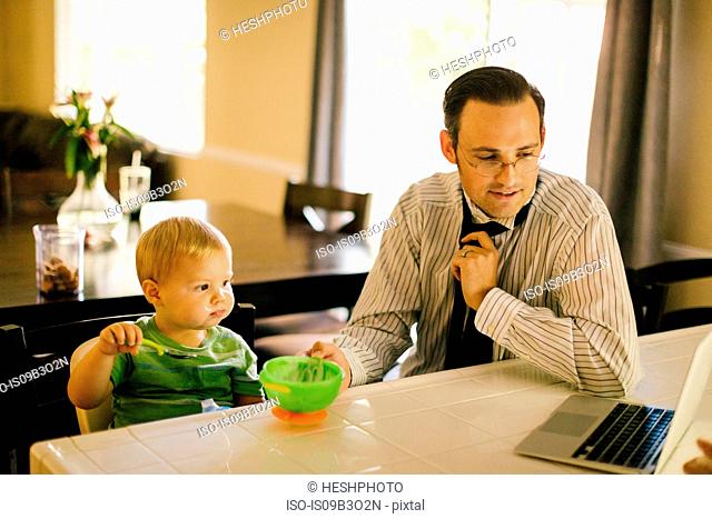 Father and young son at kitchen table, son eating breakfast, father putting on neck tie, looking at laptop