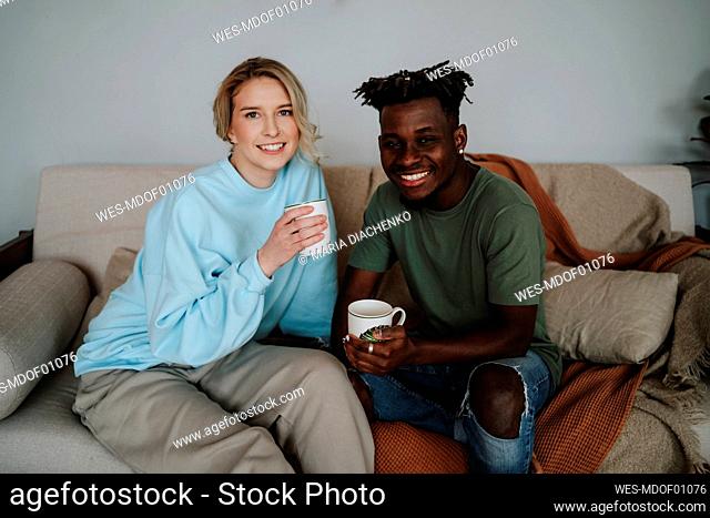 Smiling couple holding coffee mugs sitting on sofa at home