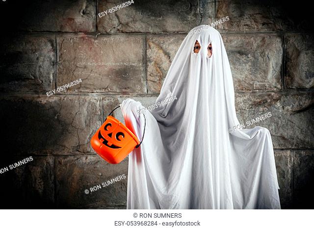 Young boy dressed in a bed sheet to be a ghost, and holding a pumpkin pail for halloween