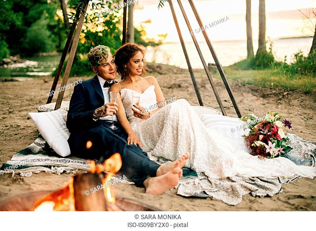Bride and groom on picnic blanket by lakeside campfire, Lake Ontario, Toronto, Canada