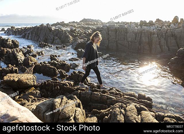 Teenage girl walking across jagged rocks, exploring rock pools by the ocean