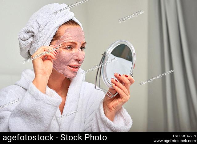Attractive woman wearing a face mask beauty treatment holding a mirror as she plucks her eyebrows with tweezers