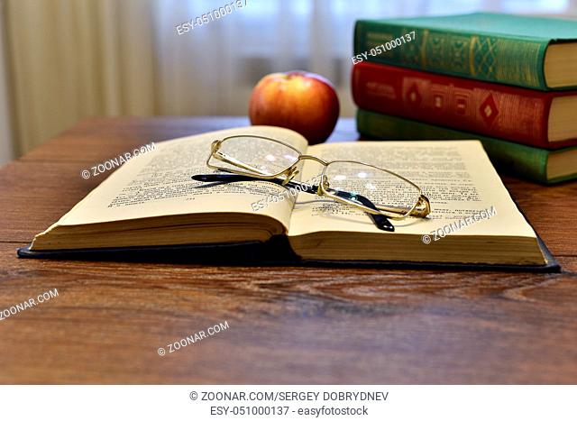 Open book with glasses, apple and stack of books on the table