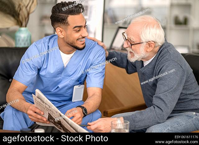 Old man in eyeglasses patting his smiling cheerful Caucasian in-home male caregiver on the shoulder