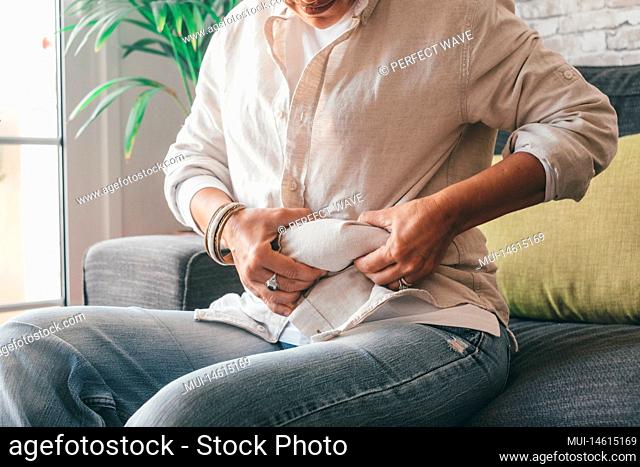 Midsection of woman hands holding her belly fat sitting on sofa at home. Caucasian lady grabbing excessive fat on her abdomen