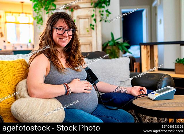 Happy portrait of a young woman in later stages of pregnancy carrying out routine health checks at home, wearing a blood pressure meter with arm cuff