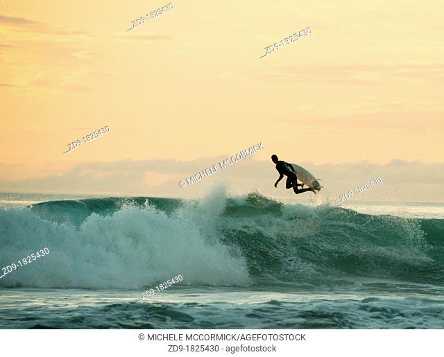 A surfer leaps back over a breaking wave in the peach-hued sunset
