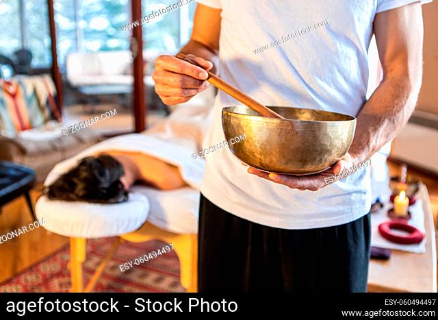 Massage therapist standing with a singing bowl in hands and giving a sound massage, to his client lying on the massage bed at the background