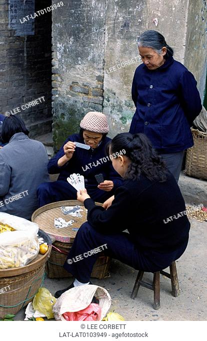 Town street. Three women playing cards at low table. Dark blue clothing. Shopping bags