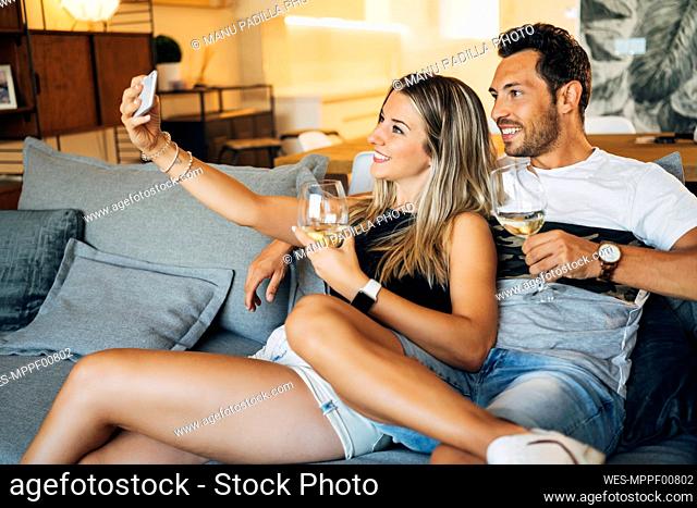 Happy couple sitting on the couch with glasses of white wine taking selfie with smartphone