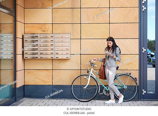 Businesswoman beside office building, walking bicycle, using smartphone