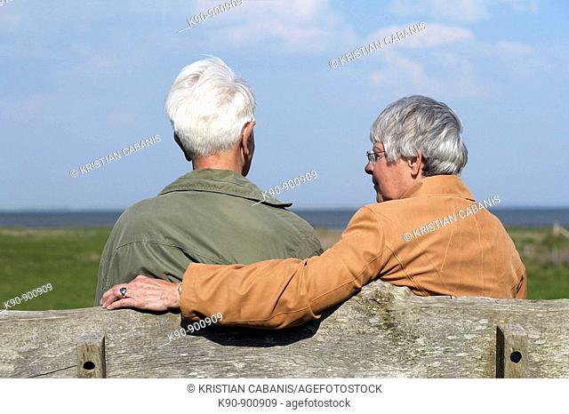 Elderly couple sitting on a bench and seen from backside while the woman is looking at the man with bright blue sky in the background, Island of Amrum