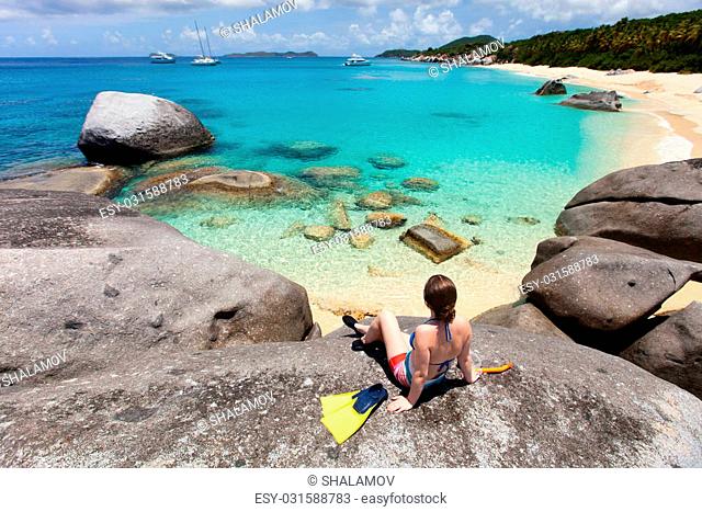 Young woman with snorkeling equipment enjoying view of a tropical beach sitting on granite boulder at Virgin Gorda, British Virgin Islands, Caribbean