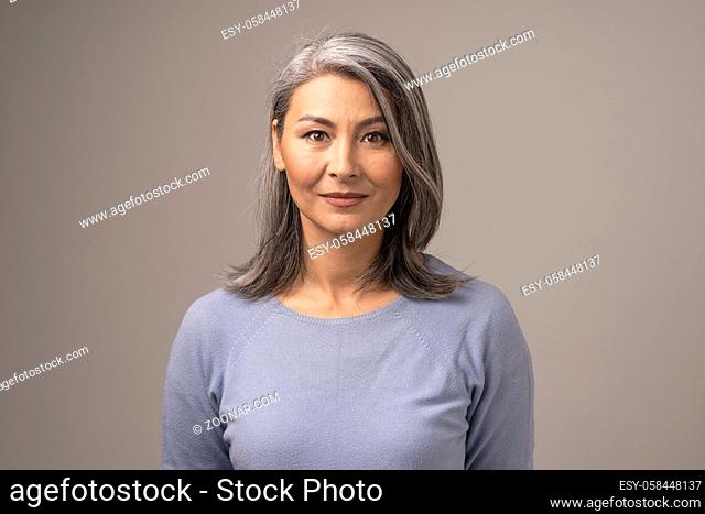 Beautiful Mongolian Woman with Gray Hair on a Gray Background. Woman's Face Has a Relaxed Smile. She is Wearing a Soft Blue Blouse. Close Up Shoot