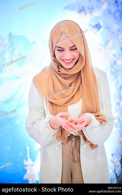 portrait of beautiful muslim woman in fashionable dress with hijab making traditional prayer to God, keeps hands in praying gesture isolated on modern winter...