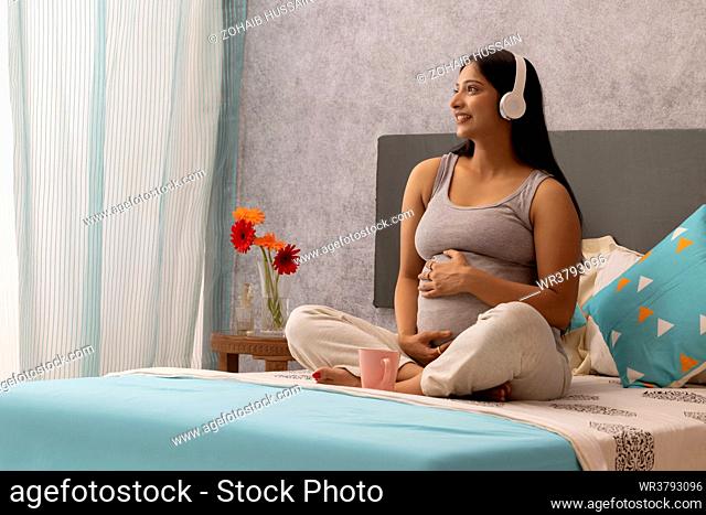 Pregnant woman listening to music with headphone while sitting on bed at home