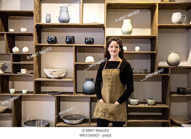 Woman with curly brown hair wearing apron standing in her pottery shop, smiling at camera