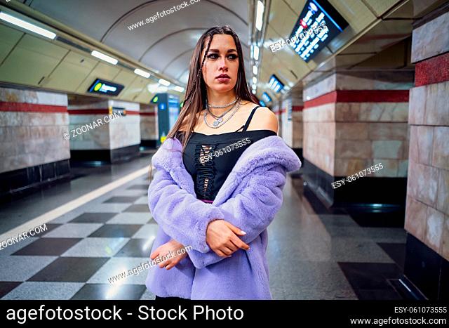 Fashion model girl in purple synthetic fur coat standing bared her shoulders in the walking subway passage platform. Female with luxury makeup