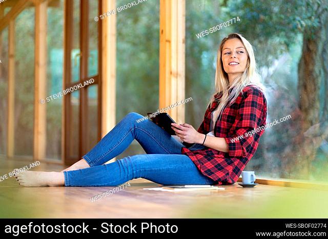 Blond woman with digital tablet looking away while sitting by window at front yard
