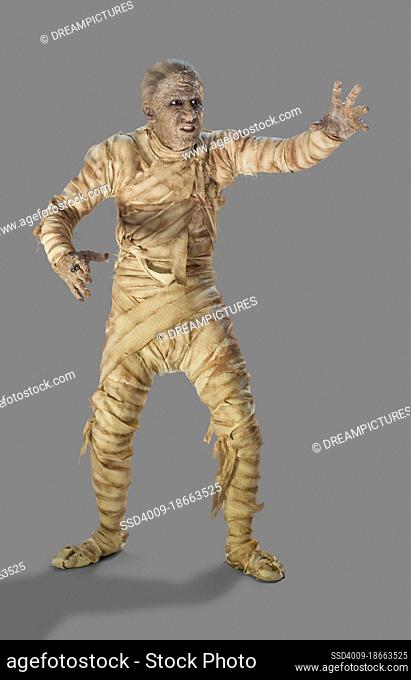 Full length portrait of a man dressed as a mummy wrapped in decaying bandages for Halloween making painful movements, against a gray background