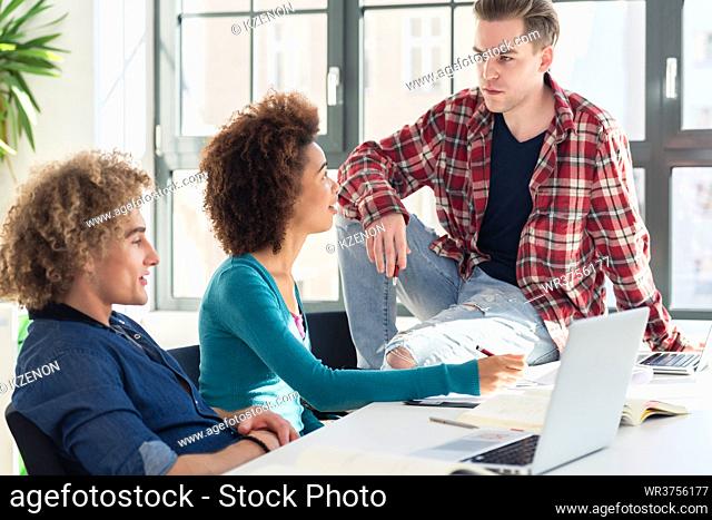 Relaxed young student sitting on desk while talking with his classmates during break in the classroom at college or university