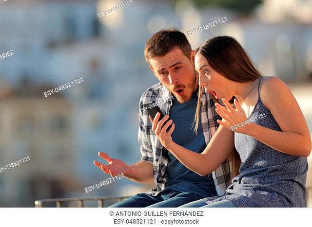 Amazed couple checking smart phone content sitting on a ledge in a town