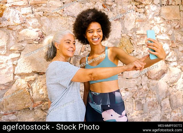 Smiling woman taking selfie with mother in front of stone wall