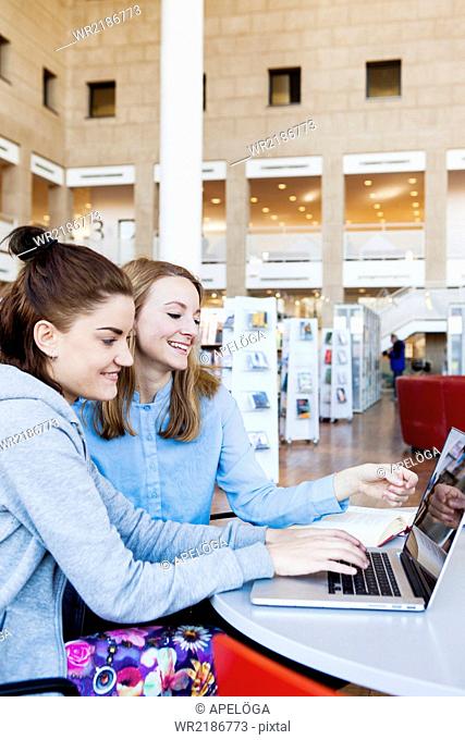 Smiling female friends with laptop and book sitting at table in library
