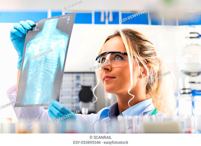 Young attractive female scientist with protective eyeglasses examiming X-ray photography film results of the human chest in the scientific hospital clinic...