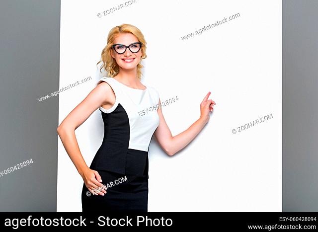 Beautiful stylish business lady in dress and glasses showing something on white wall. Blond young woman standing on grey background. Copy space
