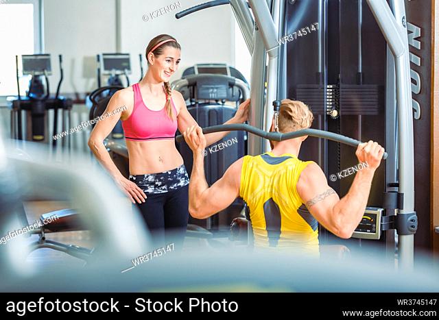 Fit beautiful woman smiling with admiration at a strong man exercising overhead lat pushdown at the cable machine in a trendy fitness club
