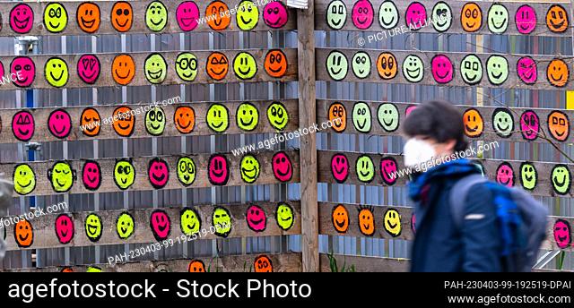 03 April 2023, Bavaria, Munich: A passerby wearing a mouth-nose covering walks past a wooden construction fence in the Museumsquartier that has been painted...
