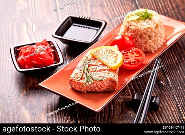 Grilled salmon fillet with rice, pickled ginger, soy sauce, tomato, lemon and herbs on dark wooden background. Healthy food