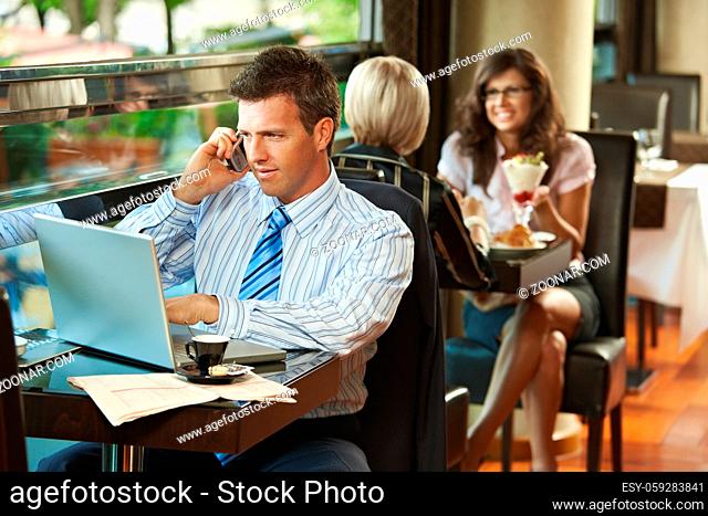 Businessman sitting at table in cafe using laptop computer, talking on mobile