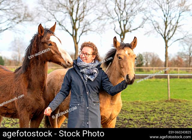 Smiling young woman in a wax coat with her 1 year old stallions in the pasture. She cuddles with them to socialize them