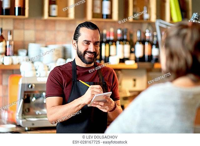 small business, people and service concept - happy man or waiter with notebook and pencil serving customer at coffee shop or bar