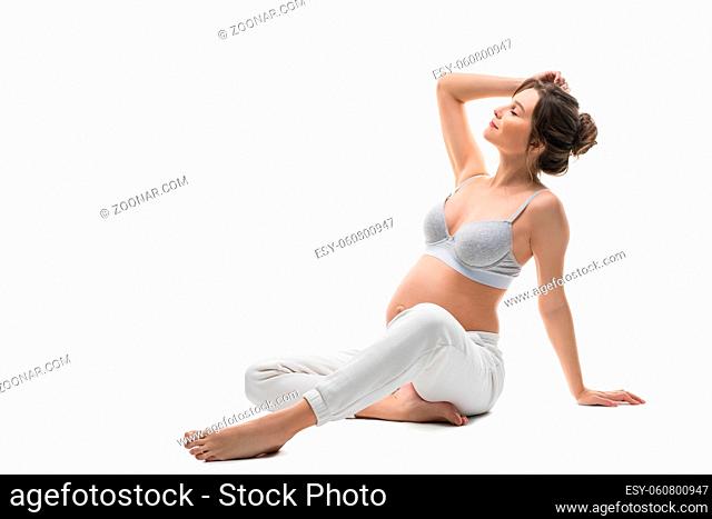 Full body side view of sensual gentle young pregnant female in white lingerie relax after stretching gracefully against white background isolated