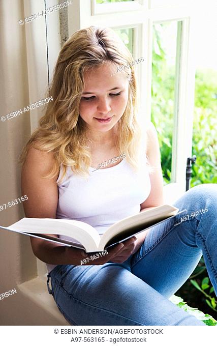 Blonde barefoot teen girl sits by an open window reading a book