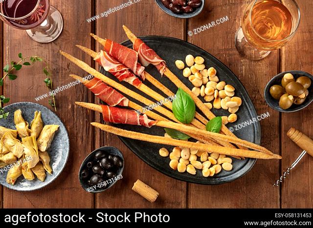 Italian antipasti. Grissini with wine glasses, parma ham and roasted almonds, with olives and artichokes, shot from above on a dark rustic wooden background