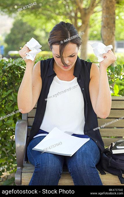 Frustrated and upset young woman with pencil and crumpled paper in her hands sitting on bench outside