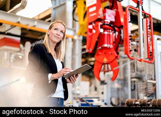 Smiling blond businesswoman looking away while holding digital tablet standing in factory