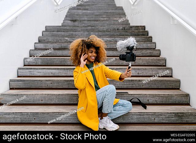 Smiling Afro woman waving hand during vlogging through camera while sitting on staircase