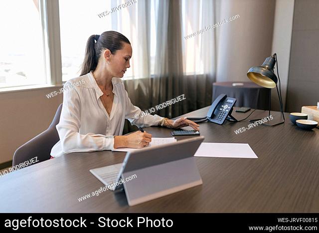 Female professional using smart phone while writing on document at desk in office