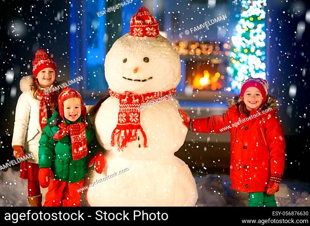 Kids build snowman on Christmas eve. Children in snowy backyard next to window to living room with Christmas tree and decorated fireplace