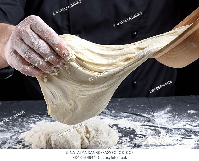 cook in black uniform kneads white wheat flour dough on a wooden table
