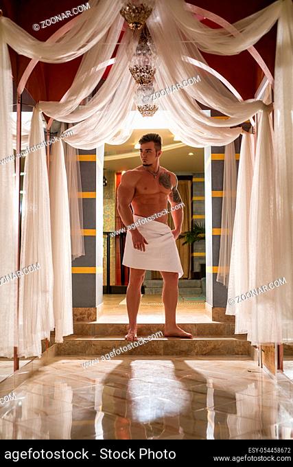 Handsome athletic tattooed man wrapped in white towel posing in luxurious sauna interior
