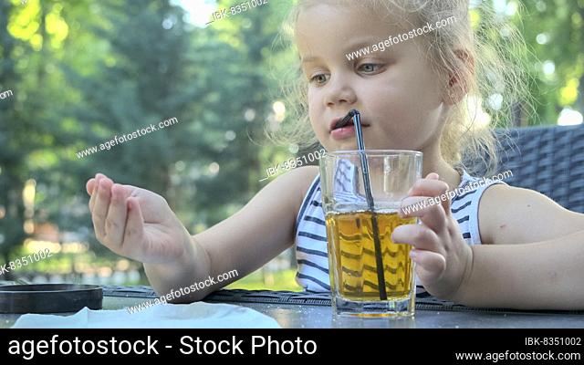Cute little girl drinks juice through straw. Close-up portrait of blonde girl drinks juice from glass through cocktail straw sitting in street cafe on the park