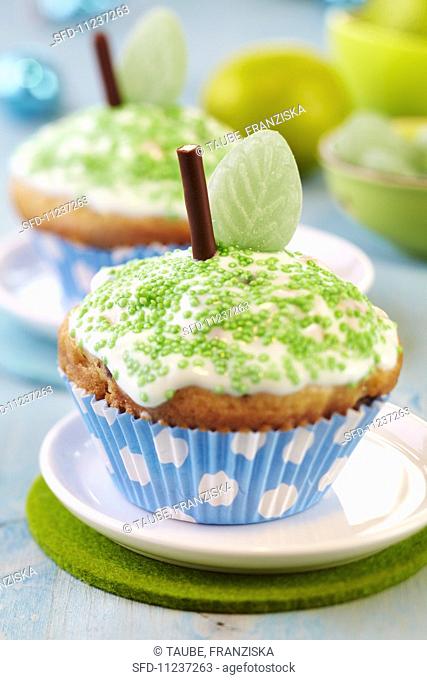 Apple muffins decorated as apples with icing, green sprinkles, chocolate sticks and leaf-shaped sweets
